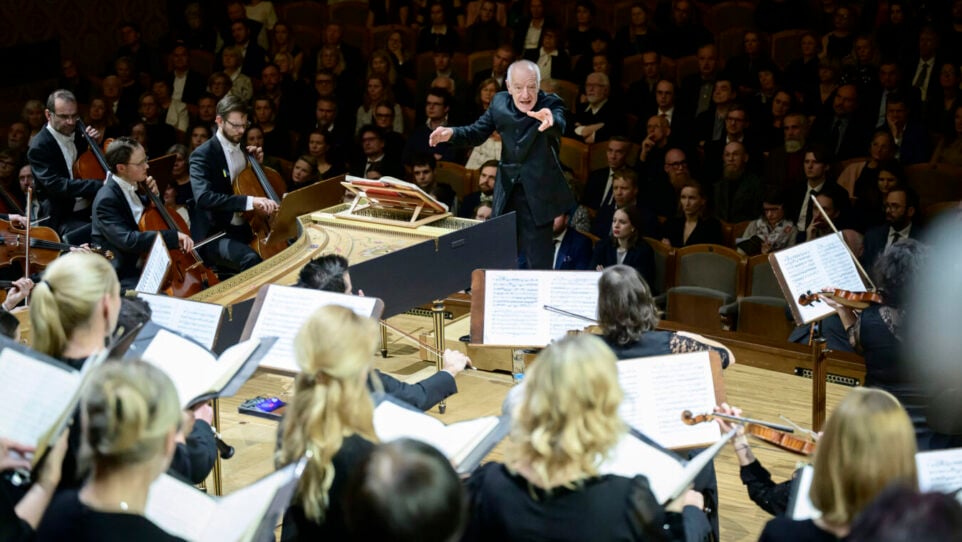 an older man conducts an orchestra with audience members behind him