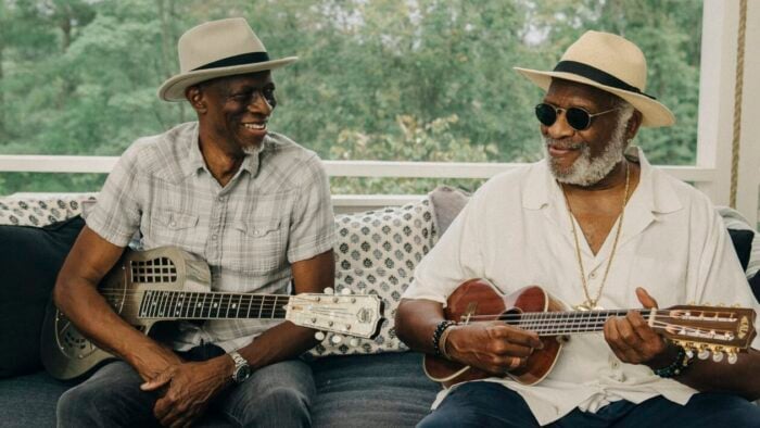 two men in hats with guitars sit smiling at one another