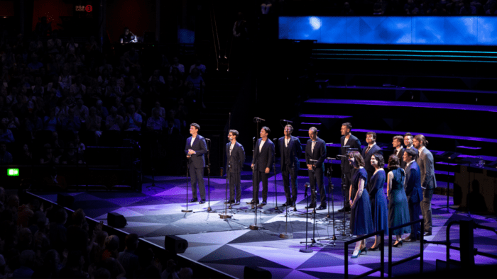 Right side view of the 14 singers of The King's Singers and VOCES8 standing in a semi-circle on stage at Royal Albert Hall; men in dark suits; women in dark blue dresses; in spotlight, audience in darkness visible in left of frame.