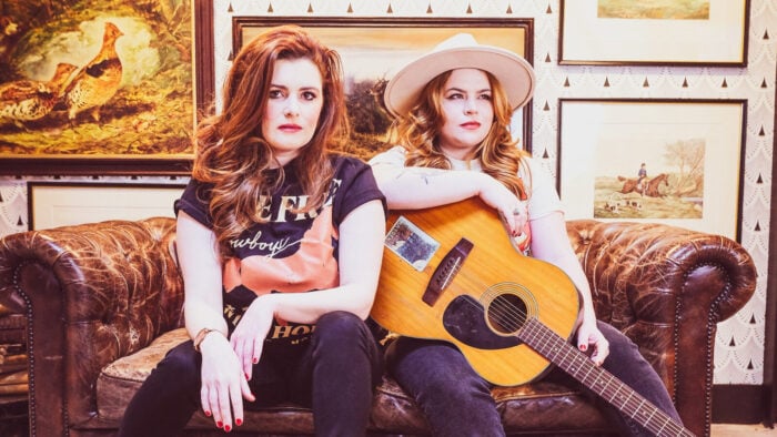 Two women, one with cowboy hat and guitar, sit on a sofa