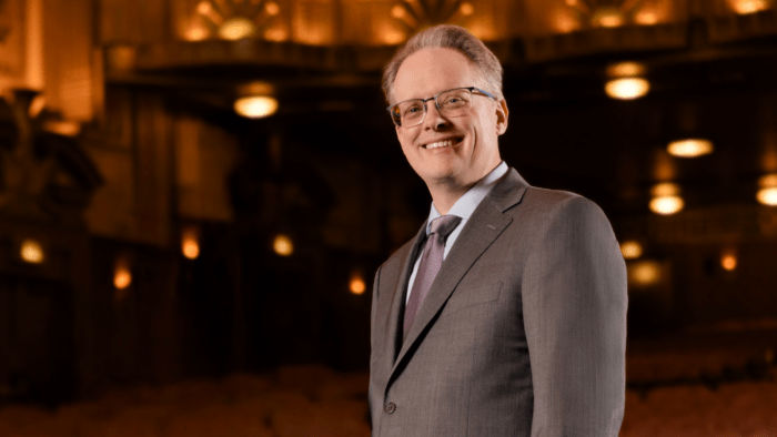 Portrait of John Mangum in a grey suit on the stage of the Ardis Krainik Theatre at Civic Opera House.