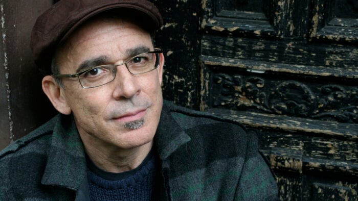 dark eyed man in glasses and a snap brim cap looks into camera