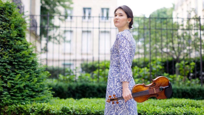 woman in blue floral dress holds violin in front of gated garden