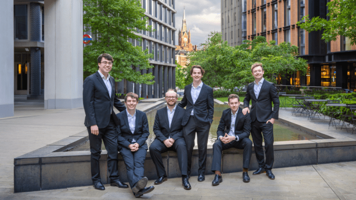 Casual portrait of The Gesualdo Six, each in a dark suit with a light shirt, no tie, in a plaza in London, St. Pancras railway station visible in background.