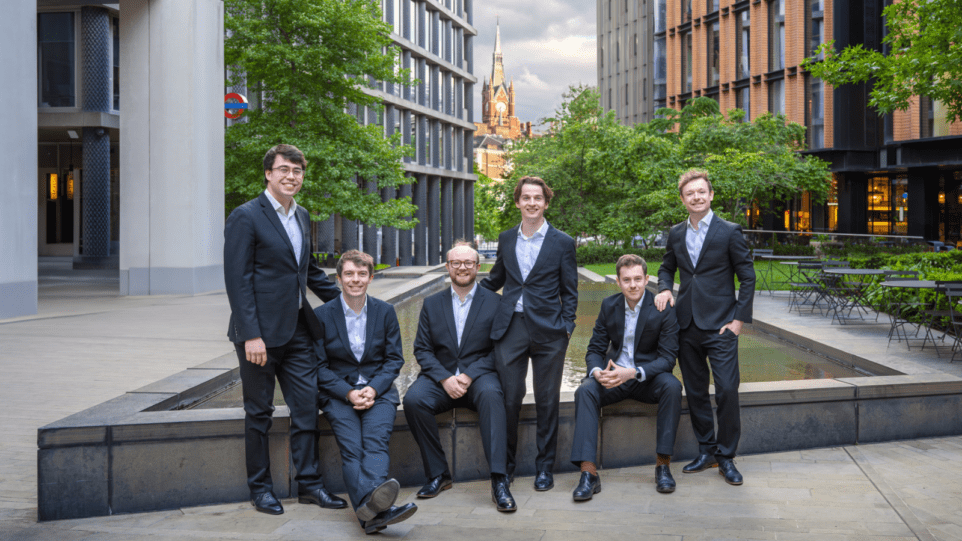 Casual portrait of The Gesualdo Six, each in a dark suit with a light shirt, no tie, in a plaza in London, St. Pancras railway station visible in background.