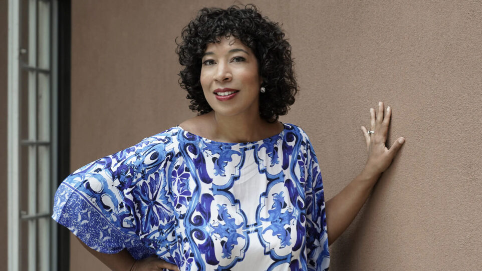 woman in blue and white patterned dress leans against brown wall