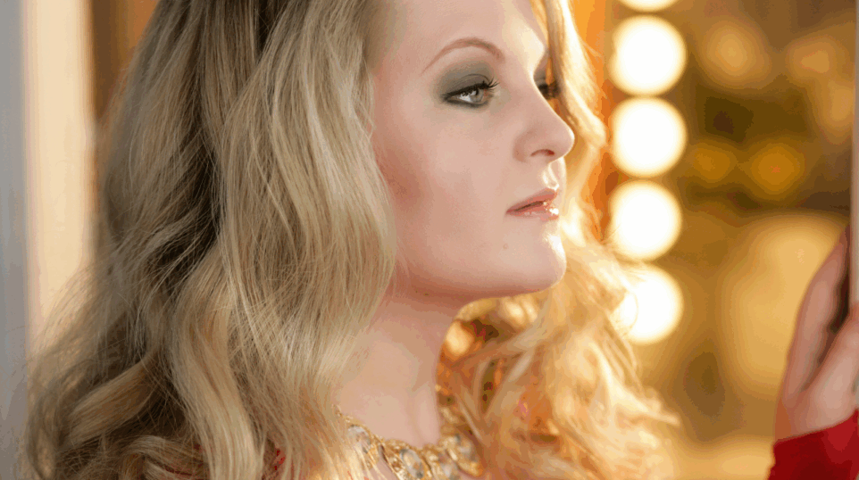 Close-up profile portrait of Elizabeth DeShong facing right in front of a dressing room mirror, its light bulbs in soft focus on her right.