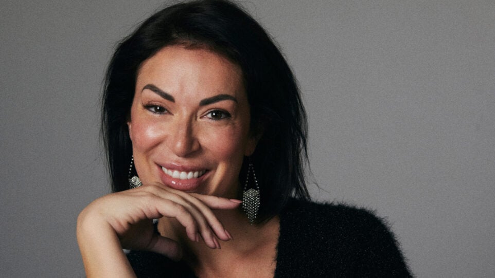 Dark haired woman with dangling earrings smiles in front of gray background