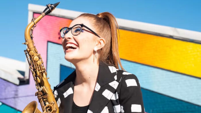 Woman smiling holding sax in front of colorful urban background and sky