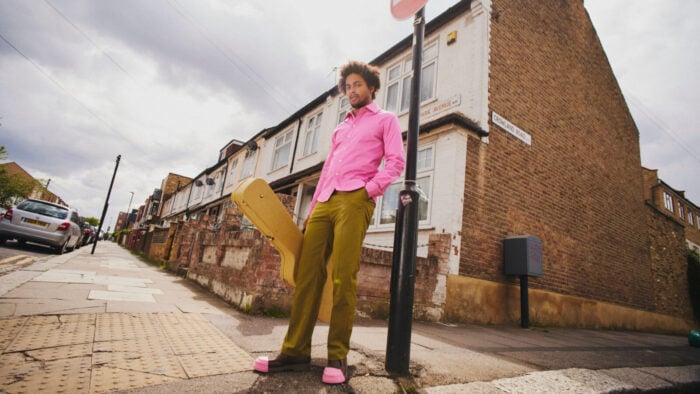 young man in bright pink shirt and shoes holds guitar case and leans against signpost in urban environment