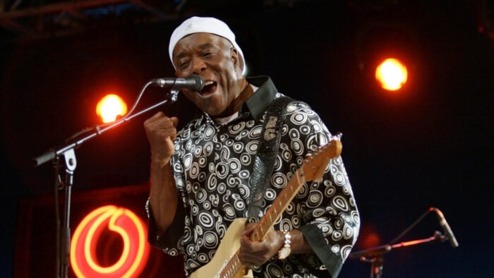Man in patterned black and white shirt and white hat plays guitar and sings onstage