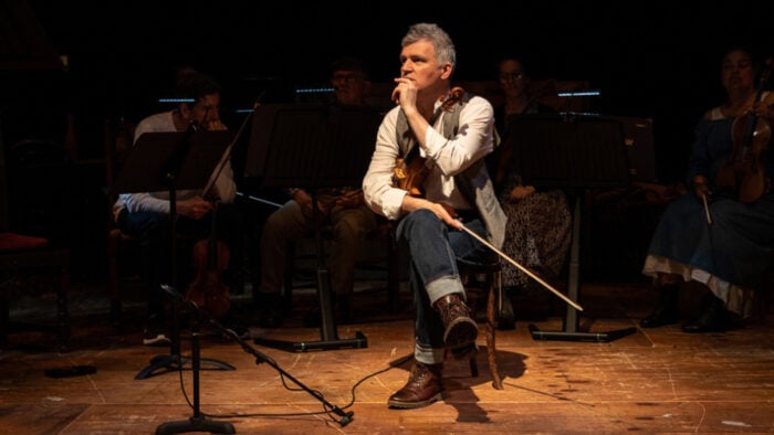 Man in jeans and a vest sits thoughtfully with violin