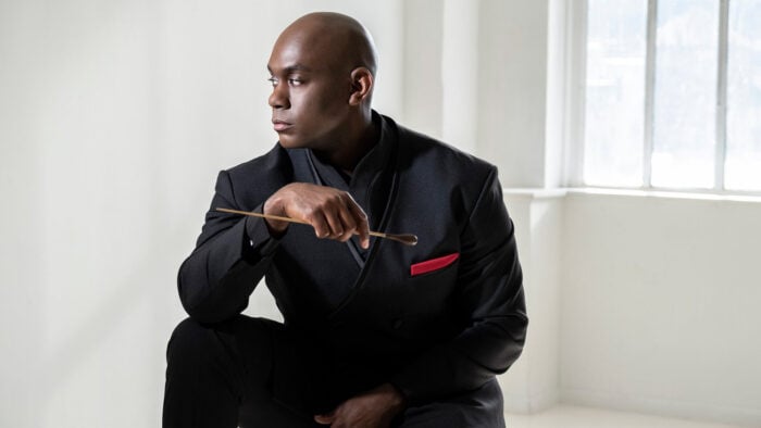 Man in black suit and red pocket square sits in white room and holds conductor's baton