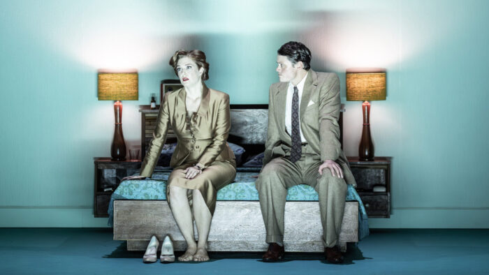 A man and a woman dressed in 1950s style seated in a bed, looking tense