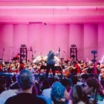 orchestra and conductor on pink-lit stage with audience looking on