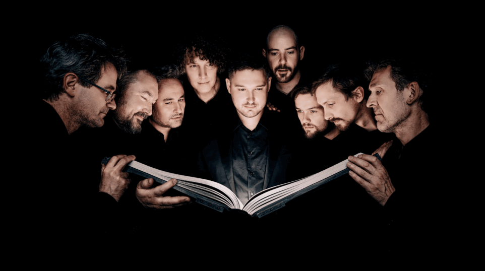 Nine men of Cappella Pratensis in dark suits huddled around one large musical score, hands of at least three of them supporting it; light source seems to be the score as all of their faces are lit from below and the background is almost pitch black.