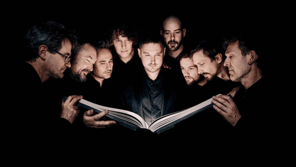 Nine men of Cappella Pratensis in dark suits huddled around one large musical score, hands of at least three of them supporting it; light source seems to be the score as all of their faces are lit from below and the background is almost pitch black.