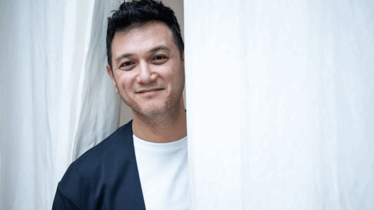 Well-lit portrait of Nicholas Phan in a white t-shirt and dark blue coat without lapels, left shoulder obscured by a white fabric, white fabric draped behind him.