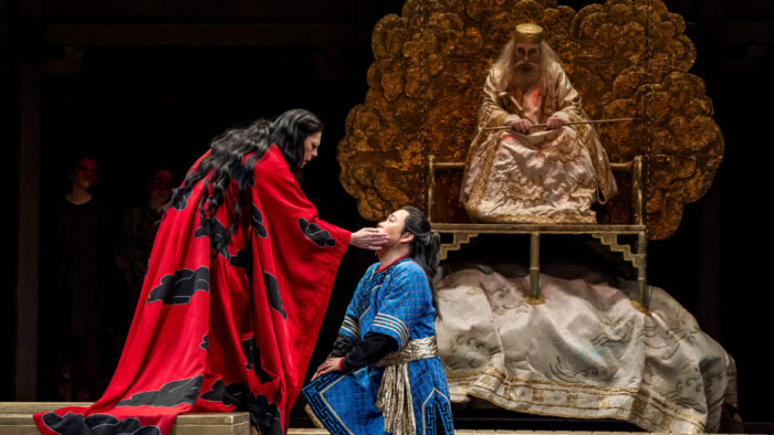 woman in vivid red robe caresses kneeling man in blue tunic, with imperial looking elderly man in background