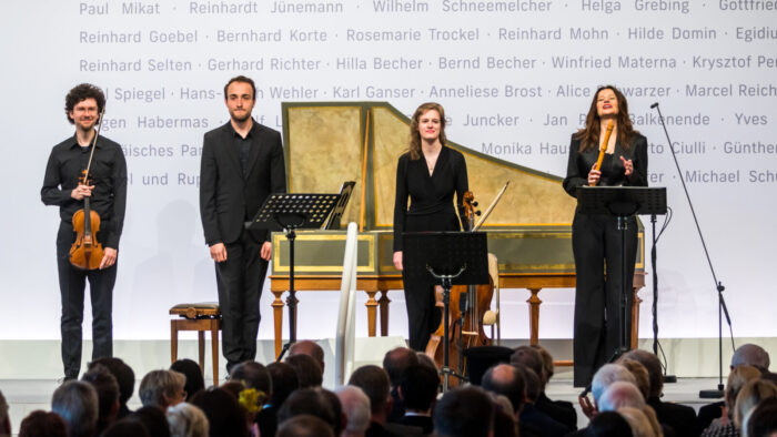 Four musicians stand with their instruments on stage in front of a harpsichord