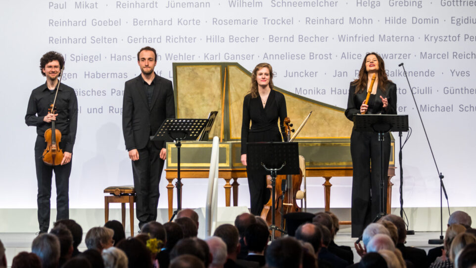 Four musicians stand with their instruments on stage in front of a harpsichord