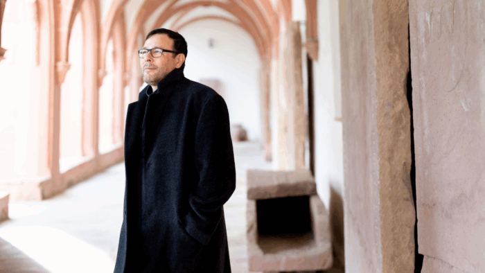 Andreas Scholl facing to the left, in dark clothing and a long black overcoat, standing in a naturally-lit arcade of (presumably) a gothic church.