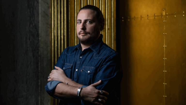 Portrait of Florian Sempey in dark denim shirt, hair pulled back in a ponytail, arms folded at his chest, two silver rings on each hand, two a silver bracelet on each wrist; low lighting, standing in front of a brass detail of a wall and copper-plated molding.