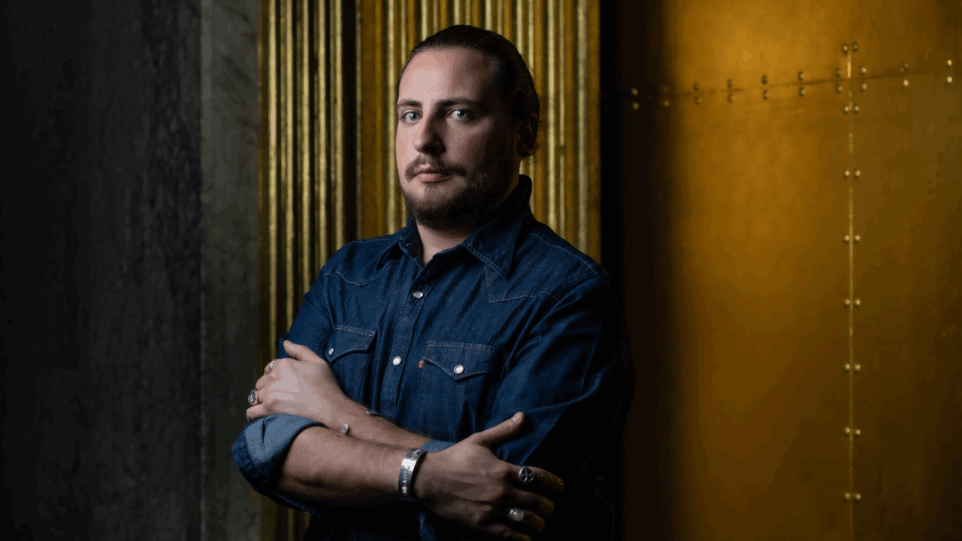 Portrait of Florian Sempey in dark denim shirt, hair pulled back in a ponytail, arms folded at his chest, two silver rings on each hand, two a silver bracelet on each wrist; low lighting, standing in front of a brass detail of a wall and copper-plated molding.