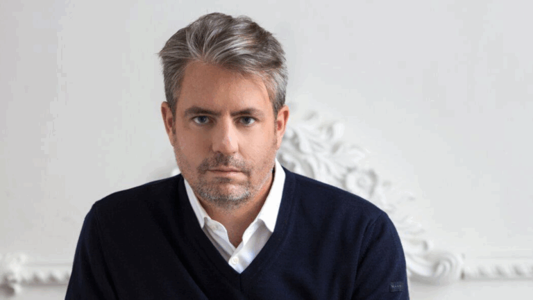 Portrait of Matthew Polenzani looking directly into camera, dark v-neck sweater over white collar shirt, white background wall has ornate molding - perhaps a laurel pattern.