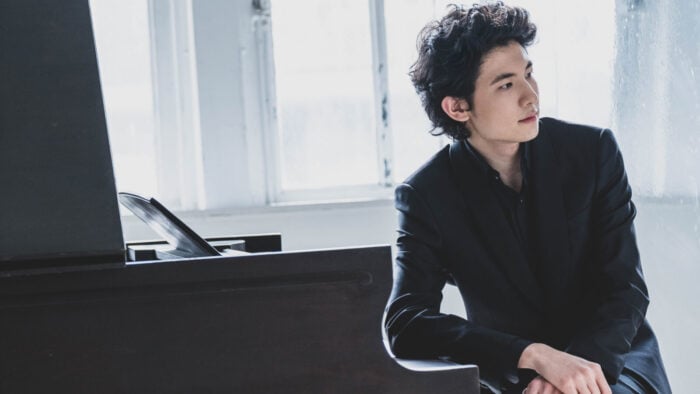 Young man, dressed darkly, looks into distance thoughtfully while leaning against a piano