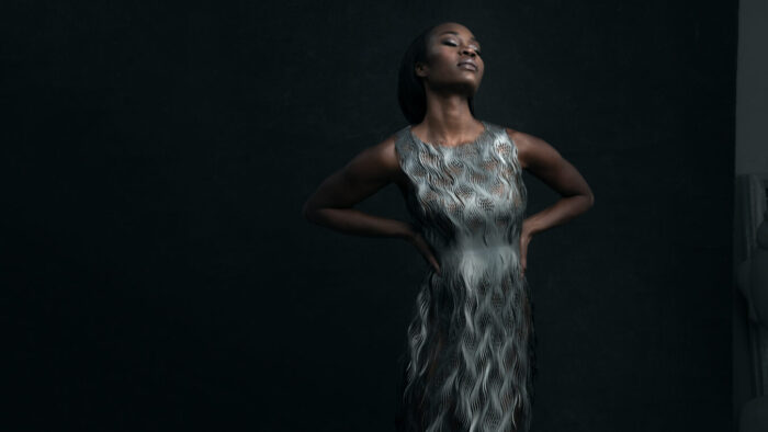 Editorial portrait of Jeanine De Bique in sleeveless translucent silver sheath dress, hands on her waist, eyes closed, chiin tilted up to expose neck; silvery makeup highlighting eyelids and lips; dark green background.