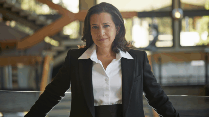 Christine Brandes in a tuxedo, no tie, leaning against a glass railing in an outdoor setting; urban architectural features out of focus in the background; natural lighting.