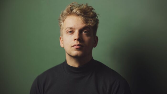 Muted portrait of Hugh Cutting in a black mock turtle neck, staring into the middle distance; green background.