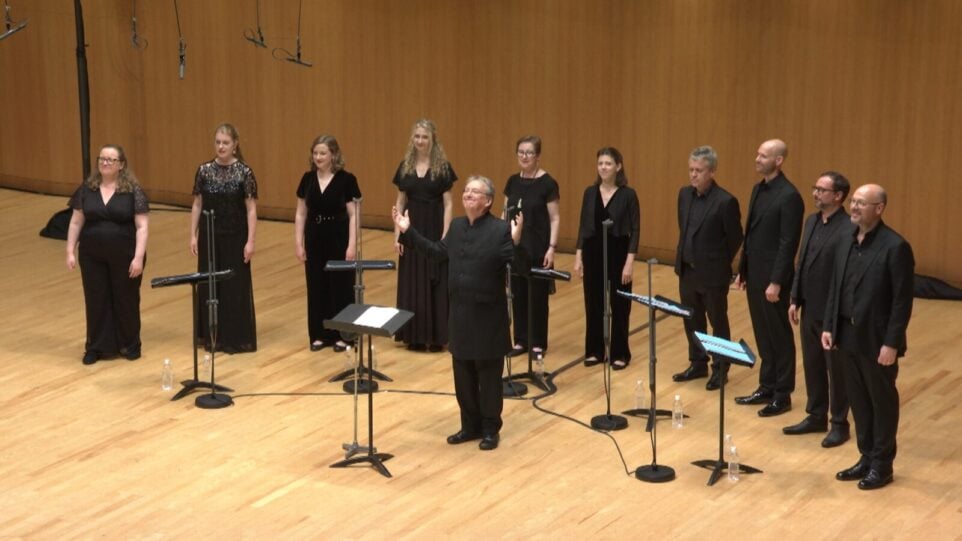 On a wooden-floored stage, left to right: dressed in concert black, six women and four men form a shallow arc behind music stands. In front is one man behind a music stand.