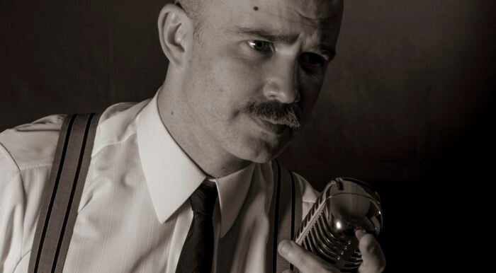 Black and white portrait of Zacahary Wilder in 50's style attire, white shirt, neck-tie, suspenders, leaning over an old-fashioned radio microphone.