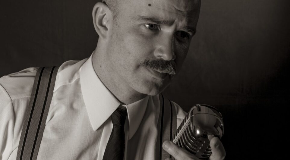 Black and white portrait of Zacahary Wilder in 50's style attire, white shirt, neck-tie, suspenders, leaning over an old-fashioned radio microphone.