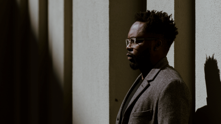 Artistic portrait in profile of Will Liverman in a grey or brown overcoat in an outdoor urban setting abstracted by vertical shadows. Liverman is in right of frame, facing directly left.