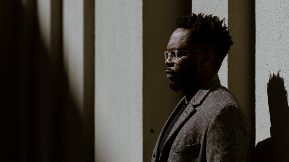 Artistic portrait in profile of Will Liverman in a grey or brown overcoat in an outdoor urban setting abstracted by vertical shadows. Liverman is in right of frame, facing directly left.