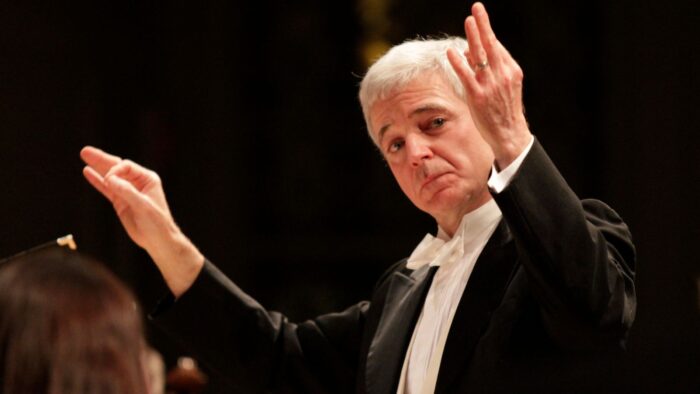 Performance image of conductor Richard Webster in black tuxedo, white bow tie; looking over his left hand, gesturing with both hands, dark background out of focus.