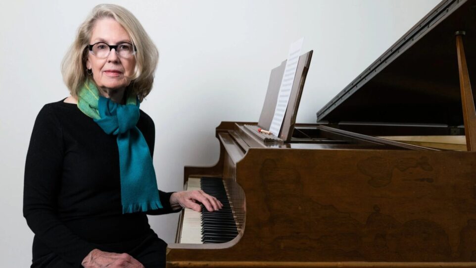An older white woman wearing dark clothes and a colorful scarf sits at her dark brown piano, hand on the keys and eyes looking at the camera.