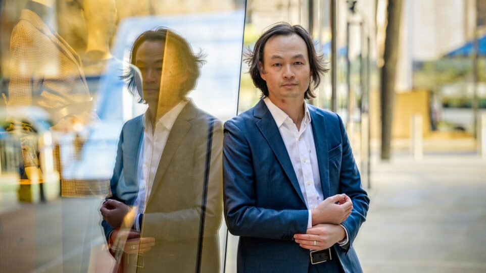 Christopher Tin in blue sit, white shirt open at collar, standing against window of a boutique creating mirror image to his left; mannequins visible behind reflection. Natural lighting.
