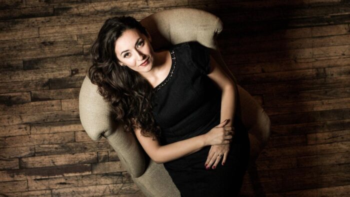 Portrait of Rosa Feola taken overhead; Feola is leaning back in a neutral colored armchair wearing a black dress; he hair and dress creating a strong contrast with the chair; and hardwood floor which serves as the background.