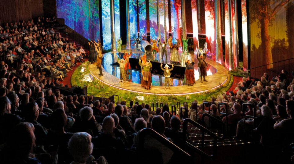 Performance image of "The Cunning Little Vixen" in the Pote Theatre at the Blank Performing Arts Center at Simpson College: Capacity audience watching well-lit set in colorful, rustic costumes; digital projections of a forest setting.