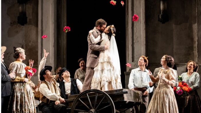 A couple standing on a wheelbarrow kissing, woman in wedding dress, man in wool suit; working-class crowd watching and cheering.