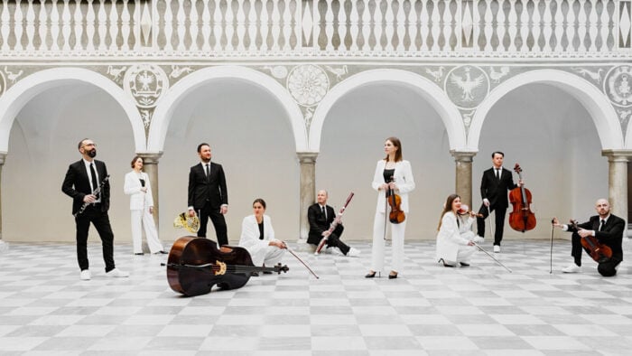 Nine musicians of ensemble minui (five string players and four wind players) in a Baroque architectural setting, checkered marble floors and ornate columns, natural lighting.