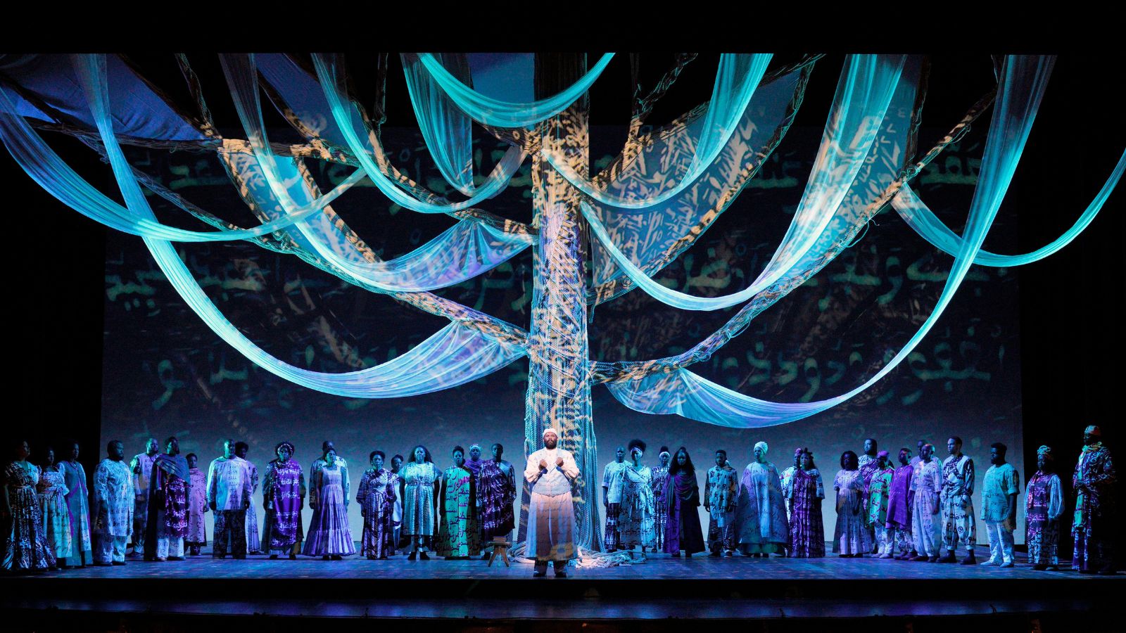 Production image from "Omar;" mixed chorus in African-inspired costume, richly patterned, mostly in puprple and white; soloist in white in center if image; fabric draped from ceiling creating upside down arch patterns; patterns similar to costumes projected onto those arches; in sombre blue light with soloist in white spotlight.