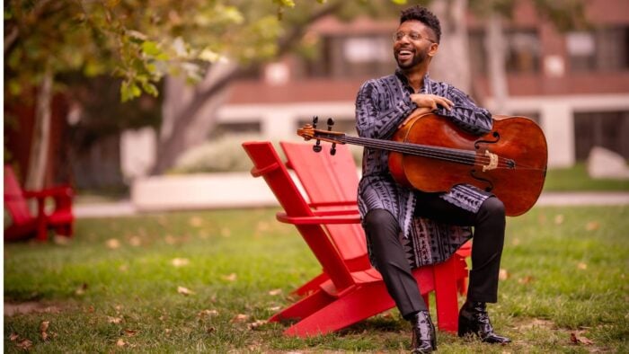 Seth Parker Woods in an urban park, seated on the arm of a rad park bench, cello on his lap, smiling