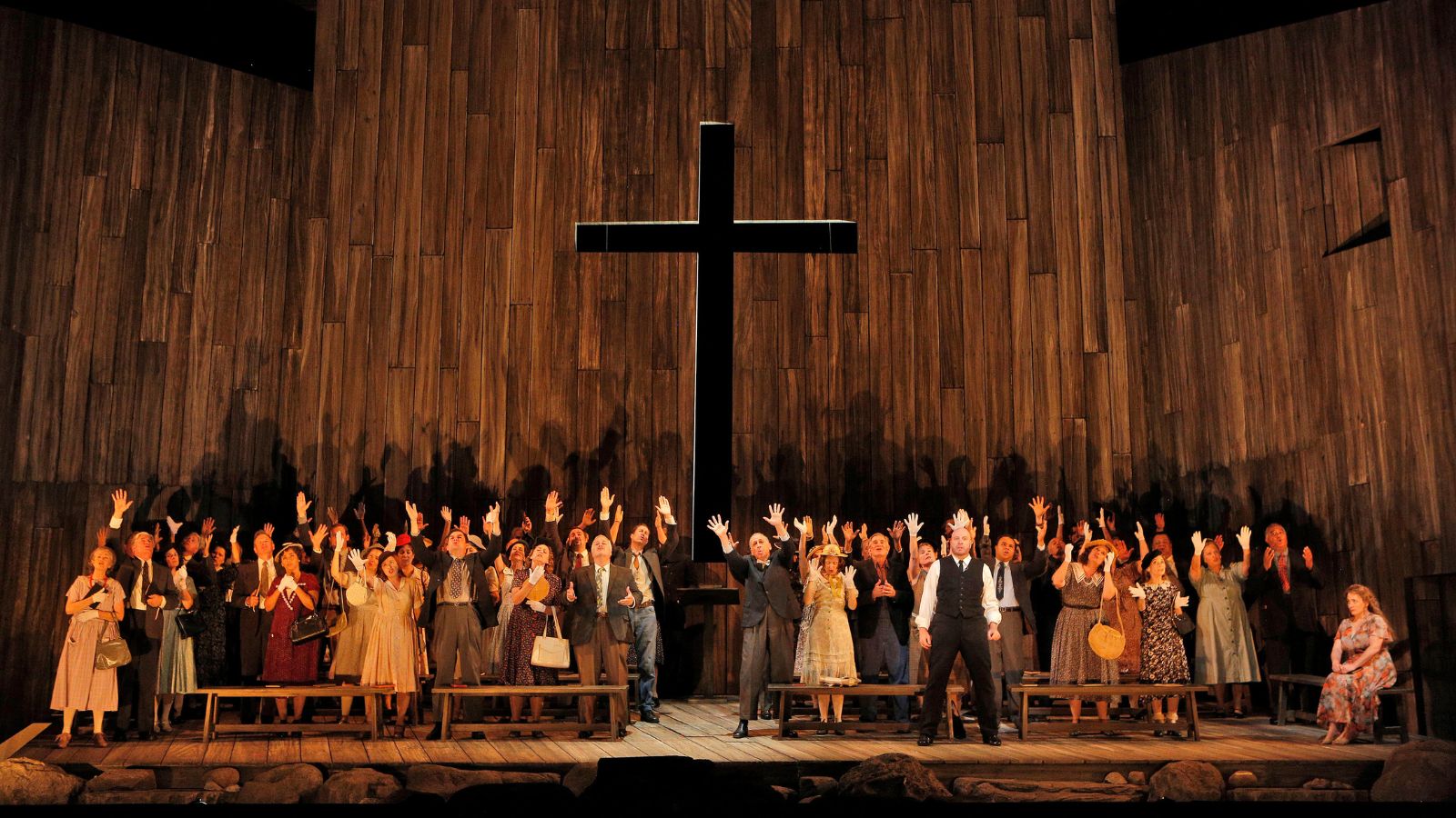 Susannah performance image; church revival meeting in wooden church with large black cross in center; congregation's hands raised in praise.