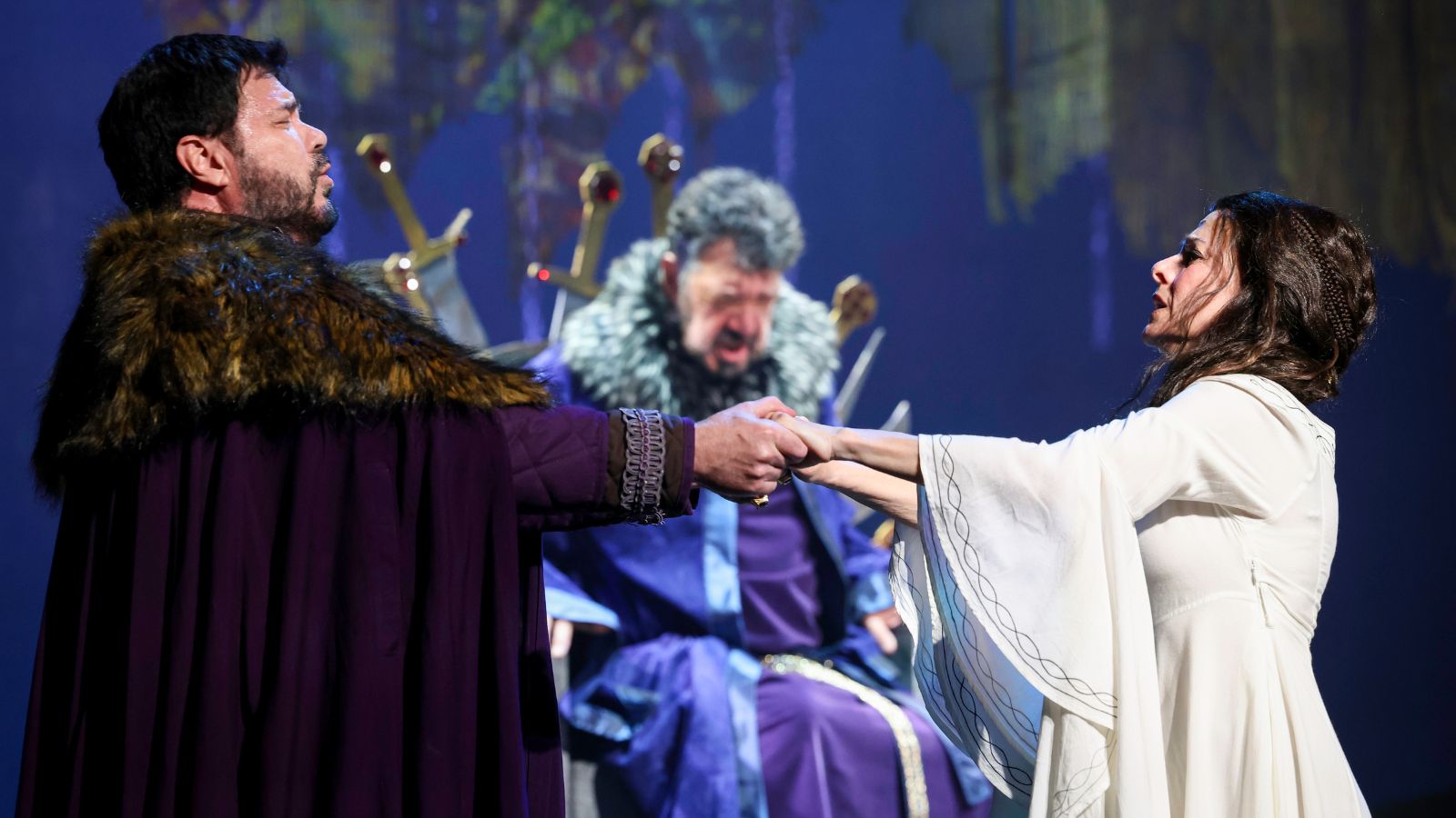 Production image of "Lamore dei tre re." Franco Pomponi clapsong hands with Maria Kanyova at arms length; Andrea Silvestrelli slumped in a throne out-of-focus in the background.