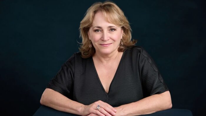 Portrait of Patricia Racette in a black blouse with elbow-lenght sleeves, looking directly into camera, hands folded in front of her; dark blue-green backdrop creating strong contrast .